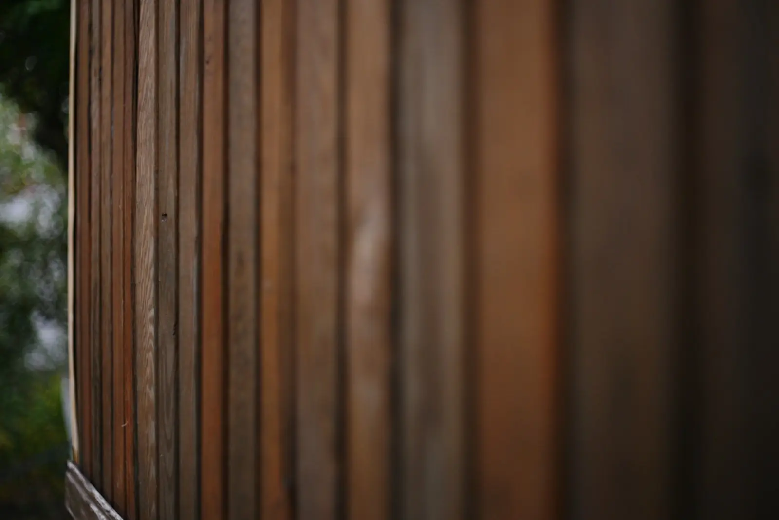 Close-up of vertical textured wooden siding, softly blurred with muted daylight and out-of-focus green foliage in the background