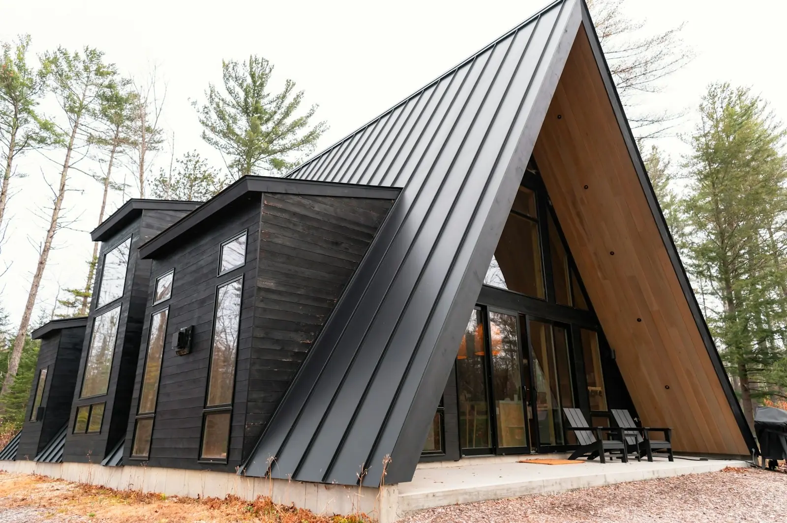 Modern black A-frame cabin with a striking standing-seam metal roof, dark wood siding, and a concrete patio with two black Adirondack chairs in a bare-tree woodland