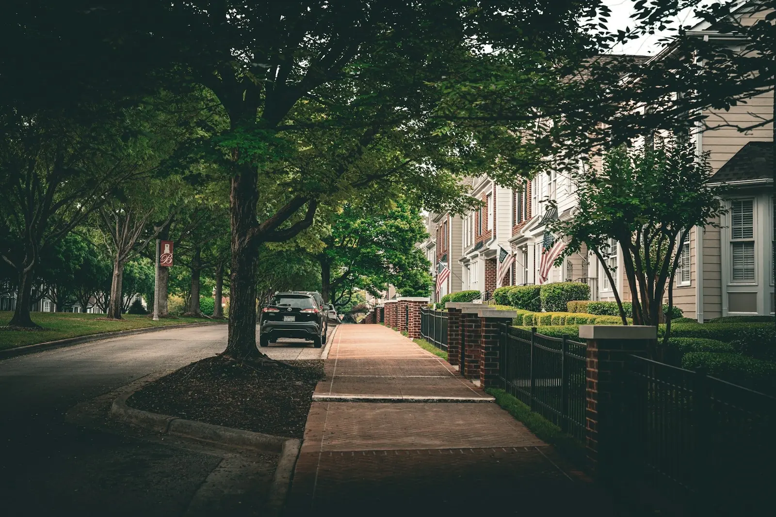 Tree-lined residential street and brick-accented sidewalk on an overcast day, with a row of traditional multi-story townhomes behind manicured hedges and an American flag, a dark SUV parked at the curb