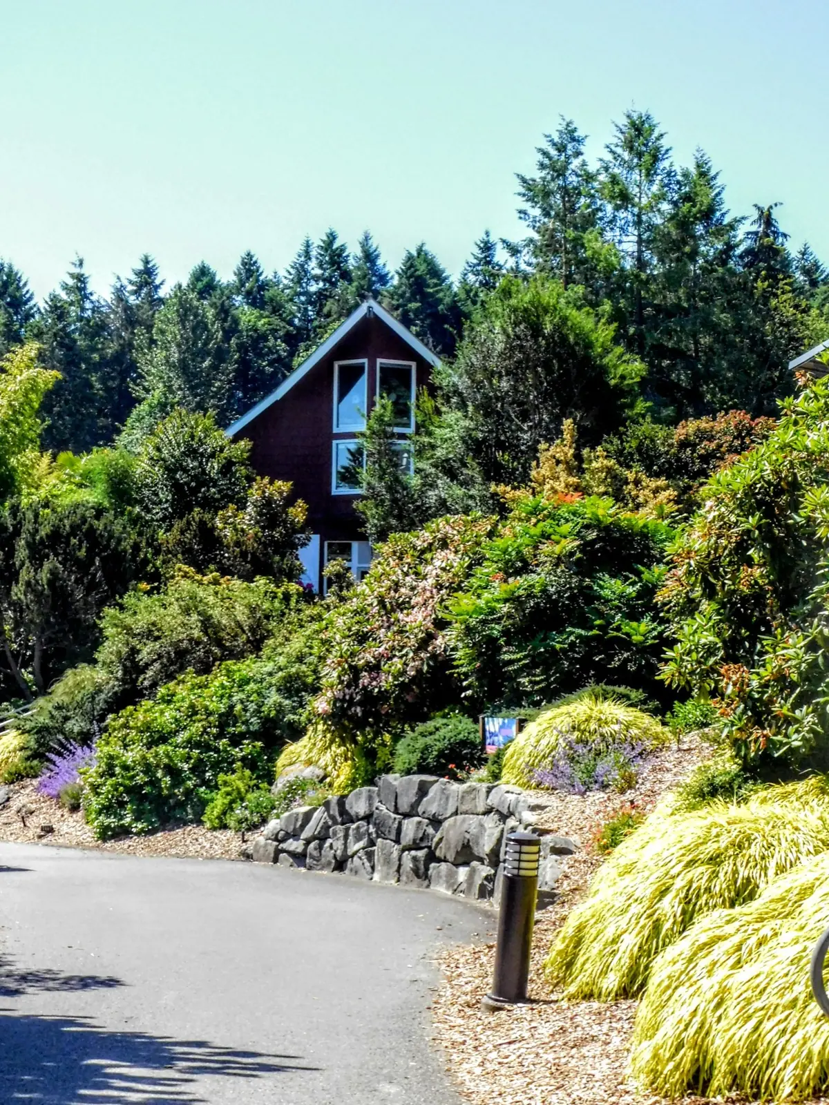Steep-gabled dark reddish-brown A-frame home nestled in a lushly forested hillside, with a curving stone-walled driveway and dense landscaping
