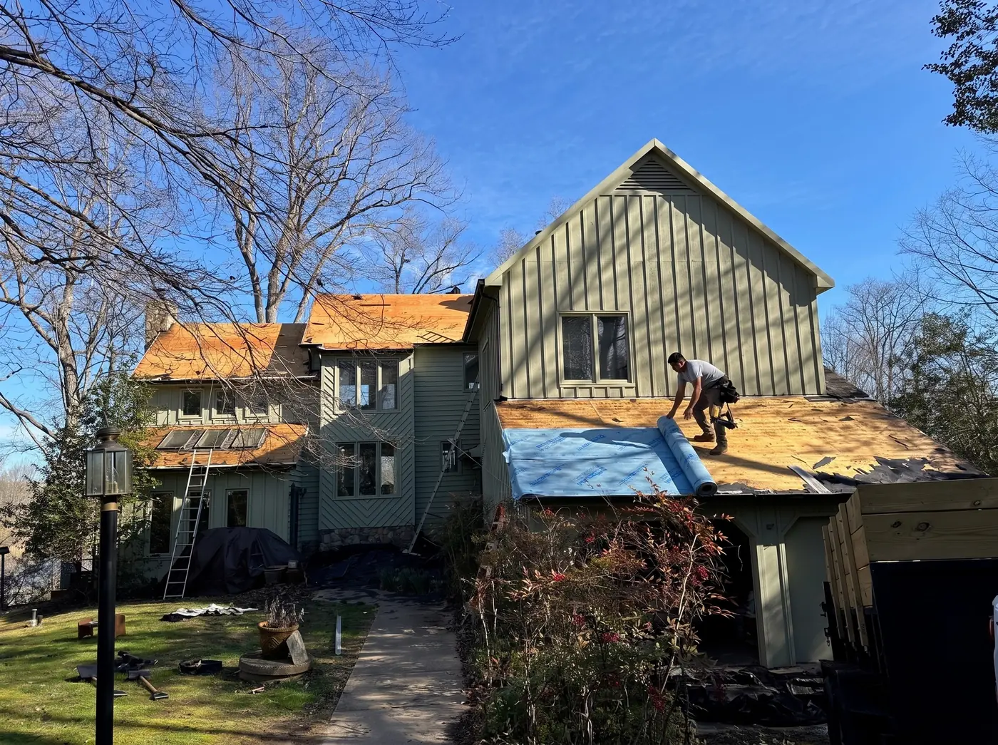 A roofer on a lower roof section unrolling blue synthetic underlayment over exposed plywood decking, with ladders and shingle bundles staged around the work area