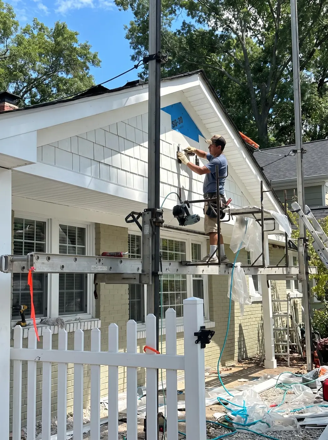 A worker on pump-jack scaffolding installing white shake siding on a residential gable, with pneumatic hoses and drop cloths behind a white picket fence