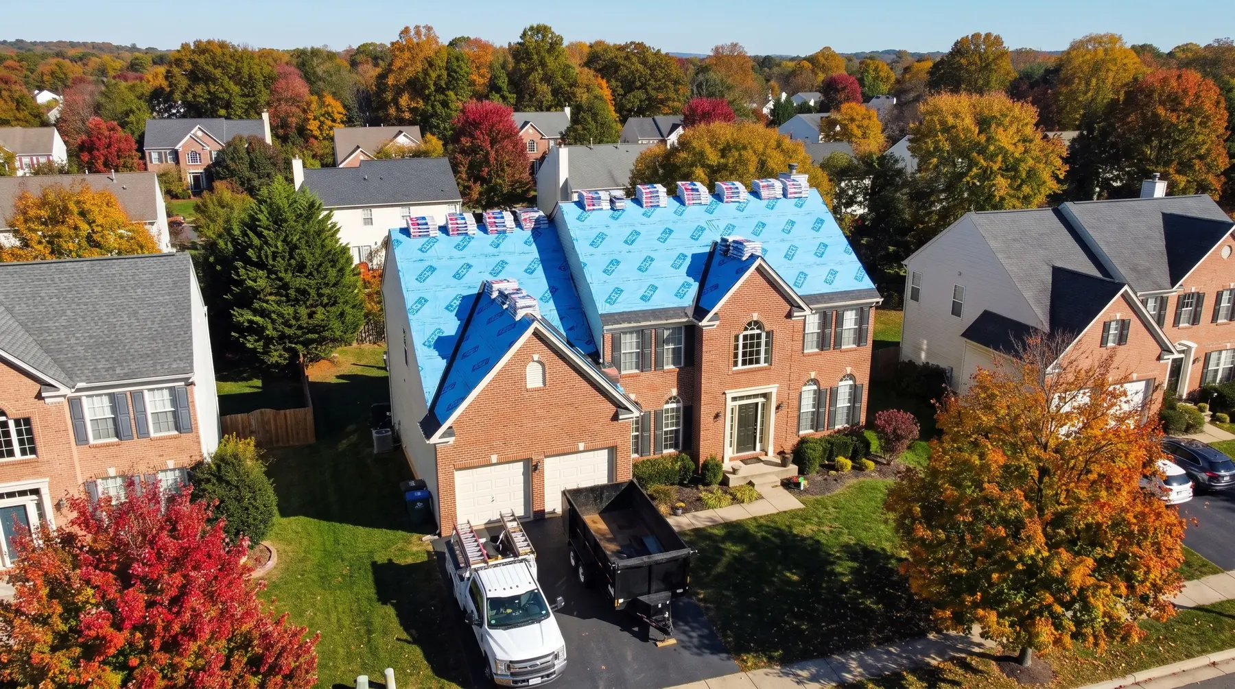 Autumn drone view of a brick colonial mid-replacement: blue underlayment down, shingle bundles staged, LCR truck below